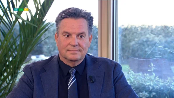 Een man in een blauw pak met stropdas zit binnen voor een raam. Links een groene plant. RTL-logo in de hoek.