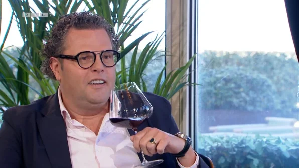 Een man met bril en krullend haar houdt een glas rode wijn vast, zittend voor een raam met groene planten op de achtergrond.