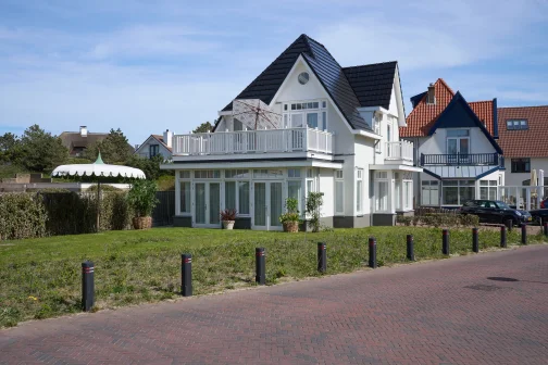 Wit huis met zwarte dakpannen, omgeven door tuin en parasol, naast andere huizen op een zonnige dag.