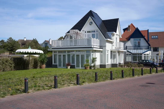Wit huis met zwarte dakpannen, omgeven door tuin en parasol, naast andere huizen op een zonnige dag.