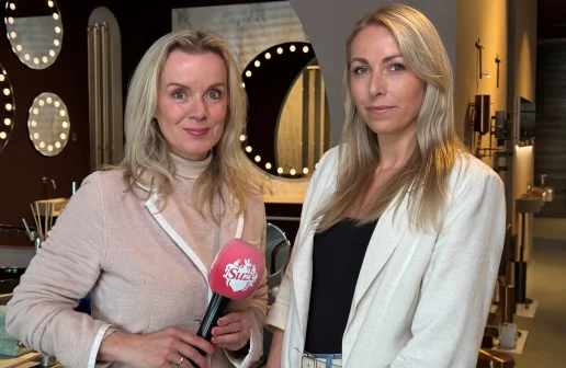Twee vrouwen in een showroom met spiegels en lampen, een van hen houdt een roze microfoon vast.