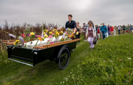 Een groep mensen volgt een kar met bloemen en een witte kist over een grasveld onder een bewolkte hemel.