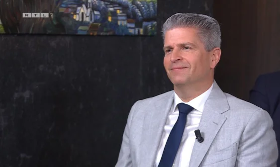 Een man in een grijs pak en blauwe stropdas zit voor een donkere muur met een schilderij. Links bovenin staat "RTL 7".