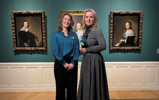 Twee vrouwen poseren voor drie klassieke portretschilderijen in een museum met groene muren en houten vloer.
