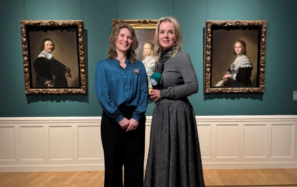 Twee vrouwen poseren voor drie klassieke portretschilderijen in een museum met groene muren en houten vloer.