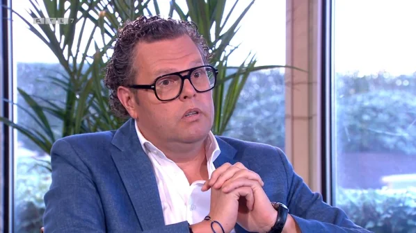 Een man met bril en blauw pak zit binnen, handen gevouwen. Achtergrond: grote ramen en planten.
