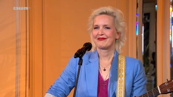 Een vrouw met blond haar speelt gitaar en zingt in een microfoon, gekleed in een blauwe blazer met een kruis ketting.