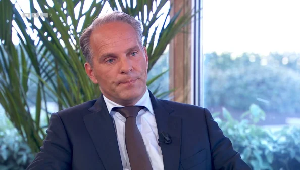 Een man in pak zit voor een raam met planten op de achtergrond. Hij kijkt serieus. Links bovenin staat "RTL 7".