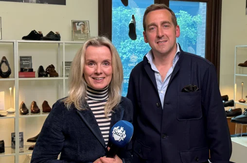 Twee mensen in een schoenenwinkel, vrouw met microfoon, man naast haar. Achter hen schoenen op planken en een raam.