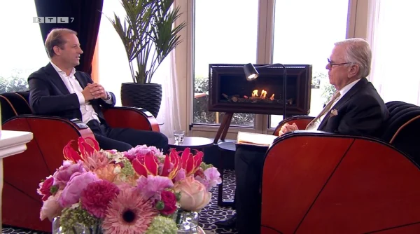 Twee mannen in gesprek in een luxe kamer met bloemen, een open haard en grote ramen.