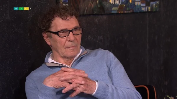 Een man met bril en krullend haar zit met gevouwen handen, draagt een lichtblauwe trui. Op de achtergrond een schilderij.