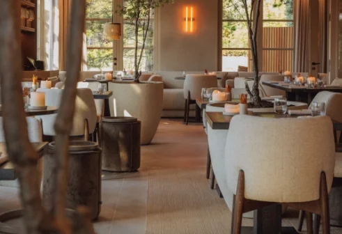 Een sfeervol restaurantinterieur met moderne, lichte stoelen, gedekte tafels, kaarsen en grote ramen met uitzicht op groen.